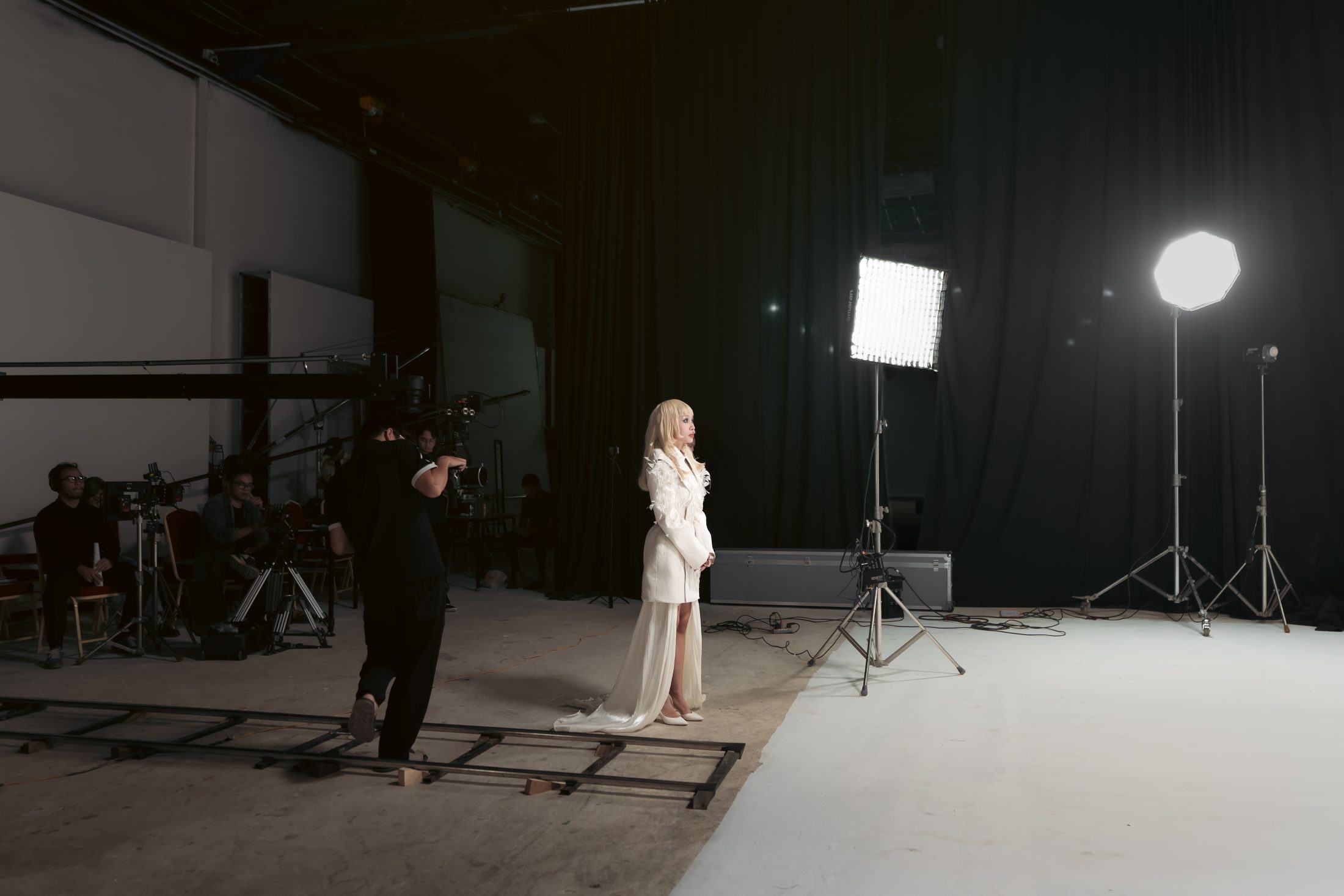 The lead actress in a white gown under studio lights, crew and camera rig to the left