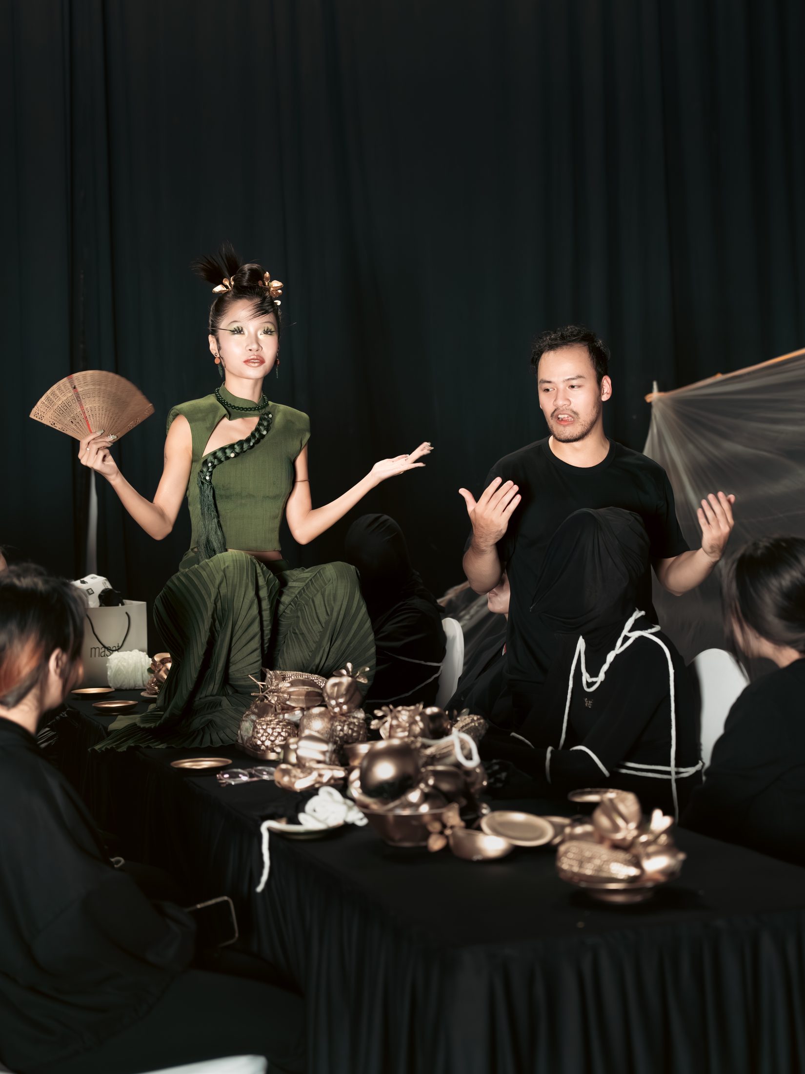 An actress in green holding a fan at a table of gilded fruit while the director gives notes